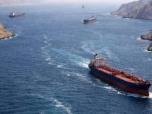 An aerial view shows several large cargo ships and oil tankers navigating through a narrow, blue waterway flanked by steep, rocky cliffs and mountains. The scene captures the strategic and rugged landscape of the Strait of Hormuz, a vital maritime corridor for global trade.