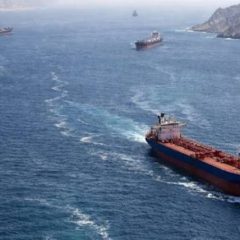 An aerial view shows several large cargo ships and oil tankers navigating through a narrow, blue waterway flanked by steep, rocky cliffs and mountains. The scene captures the strategic and rugged landscape of the Strait of Hormuz, a vital maritime corridor for global trade.