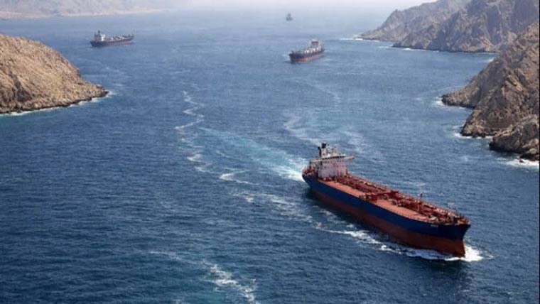 An aerial view shows several large cargo ships and oil tankers navigating through a narrow, blue waterway flanked by steep, rocky cliffs and mountains. The scene captures the strategic and rugged landscape of the Strait of Hormuz, a vital maritime corridor for global trade.