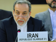 A close-up shot of Iranian Foreign Minister Abbas Araghchi, a man with grey hair, a beard, and glasses resting low on his nose. He is wearing a dark suit with a white shirt and speaking into a microphone. In front of him is a white placard reading "IRAN (REPUBLIQUE ISLAMIQUE D')".