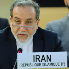 A close-up shot of Iranian Foreign Minister Abbas Araghchi, a man with grey hair, a beard, and glasses resting low on his nose. He is wearing a dark suit with a white shirt and speaking into a microphone. In front of him is a white placard reading "IRAN (REPUBLIQUE ISLAMIQUE D')".