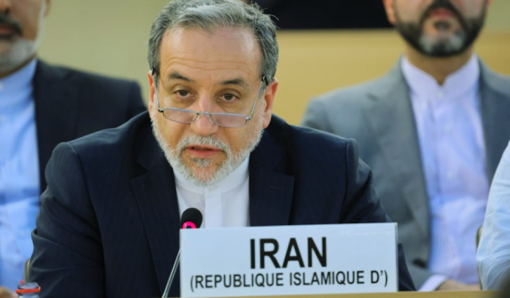 A close-up shot of Iranian Foreign Minister Abbas Araghchi, a man with grey hair, a beard, and glasses resting low on his nose. He is wearing a dark suit with a white shirt and speaking into a microphone. In front of him is a white placard reading "IRAN (REPUBLIQUE ISLAMIQUE D')".