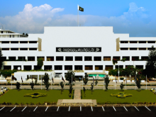 An exterior shot of the white, multi-story Parliament House building of Pakistan in 22.jpg. The national flag flies prominently at the center top. A black banner with Arabic calligraphy (the Kalima) is displayed on the building's central facade. In front of the structure, there is a neatly landscaped green lawn with small trees, flanked by an asphalt parking area with white markings under a bright, partly cloudy sky.