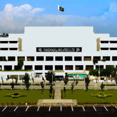 An exterior shot of the white, multi-story Parliament House building of Pakistan in 22.jpg. The national flag flies prominently at the center top. A black banner with Arabic calligraphy (the Kalima) is displayed on the building's central facade. In front of the structure, there is a neatly landscaped green lawn with small trees, flanked by an asphalt parking area with white markings under a bright, partly cloudy sky.