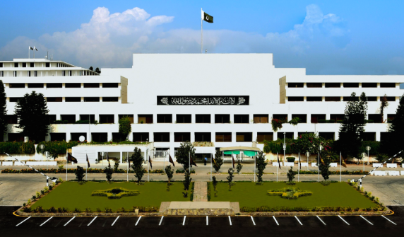 An exterior shot of the white, multi-story Parliament House building of Pakistan in 22.jpg. The national flag flies prominently at the center top. A black banner with Arabic calligraphy (the Kalima) is displayed on the building's central facade. In front of the structure, there is a neatly landscaped green lawn with small trees, flanked by an asphalt parking area with white markings under a bright, partly cloudy sky.