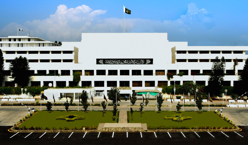 An exterior shot of the white, multi-story Parliament House building of Pakistan in 22.jpg. The national flag flies prominently at the center top. A black banner with Arabic calligraphy (the Kalima) is displayed on the building's central facade. In front of the structure, there is a neatly landscaped green lawn with small trees, flanked by an asphalt parking area with white markings under a bright, partly cloudy sky.