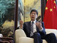 Chinese Ambassador to Iran, Kong Peiwu, wearing a suit and glasses, speaking during an interview with the Chinese national flag in the background.