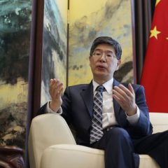 Chinese Ambassador to Iran, Kong Peiwu, wearing a suit and glasses, speaking during an interview with the Chinese national flag in the background.