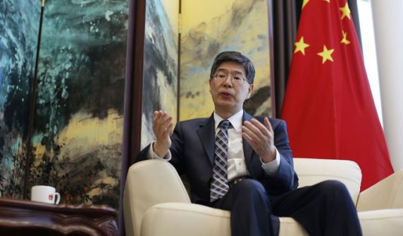 Chinese Ambassador to Iran, Kong Peiwu, wearing a suit and glasses, speaking during an interview with the Chinese national flag in the background.