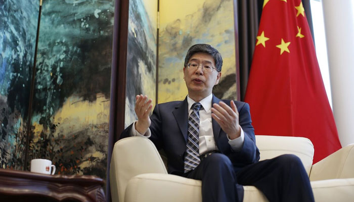 Chinese Ambassador to Iran, Kong Peiwu, wearing a suit and glasses, speaking during an interview with the Chinese national flag in the background.