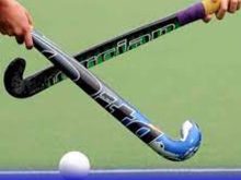 A close-up shot from the file hockey-1.jpg showing two players' hands holding field hockey sticks that are crossed in an "X" shape over a white ball on a green artificial turf.